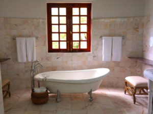 A standalone bathtub under red wood paneled windows and two towel racks in a light tile floor and walled room. 