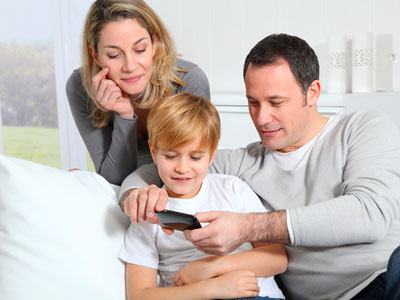 A family of three sitting and standing by a white couch looking into a black smart thermostat remote. 