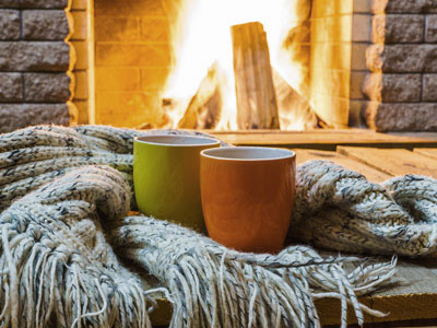 Two mugs standing next to a woolen scarf in front of a blazing fireplace. 